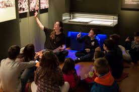 Children sitting on the floor in a museum, listening to a guide pointing at a display.